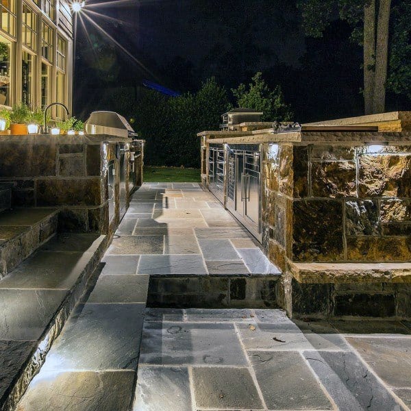 Outdoor kitchen at night with stone countertops, grill, and modern outdoor lighting