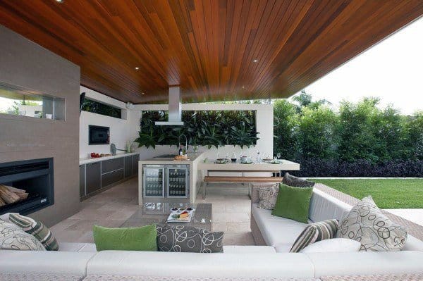 Outdoor kitchen and lounge with wooden ceiling, green accents, and lush greenery in the background