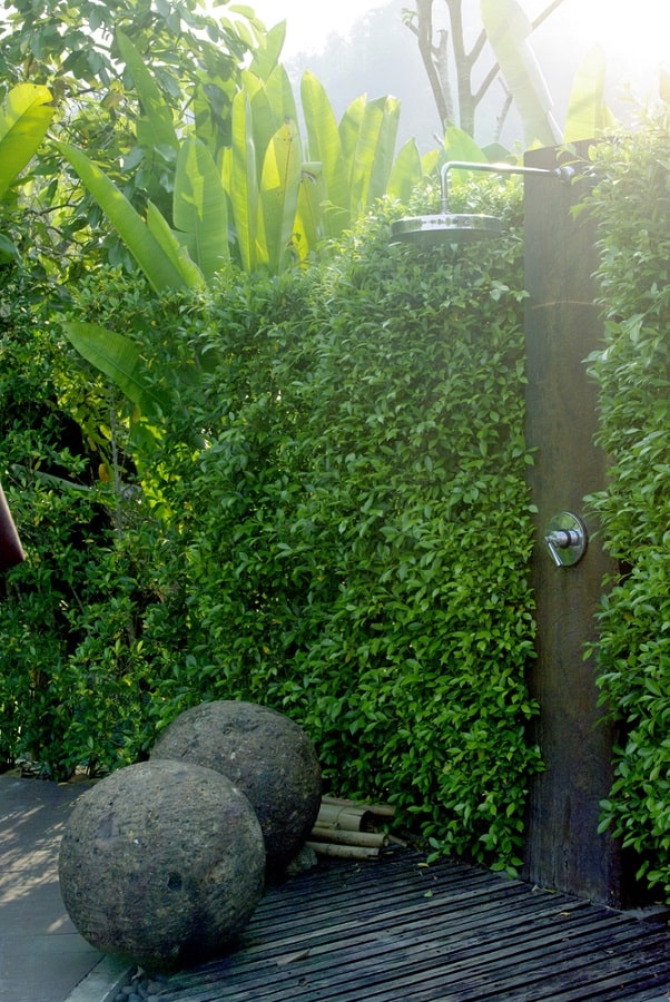 ropical outdoor shower with dense greenery, wooden floor, and decorative stone spheres.