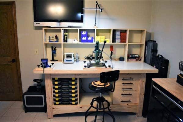 Stylish reloading bench with a mounted press, organized shelves, labeled storage drawers, and a mounted TV for convenience