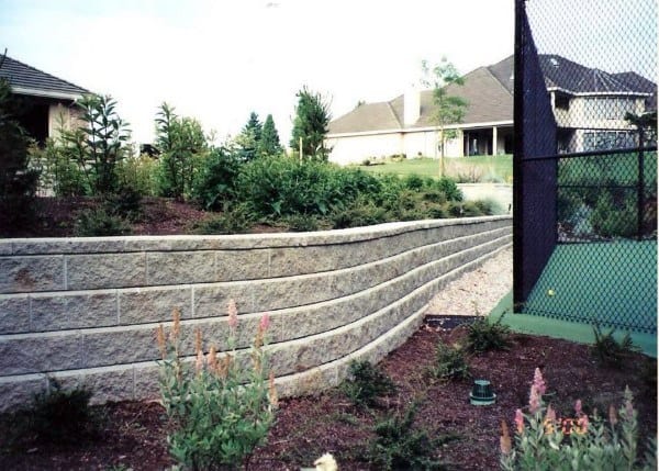 Tiered concrete block retaining walls with landscaped greenery near a tennis court.