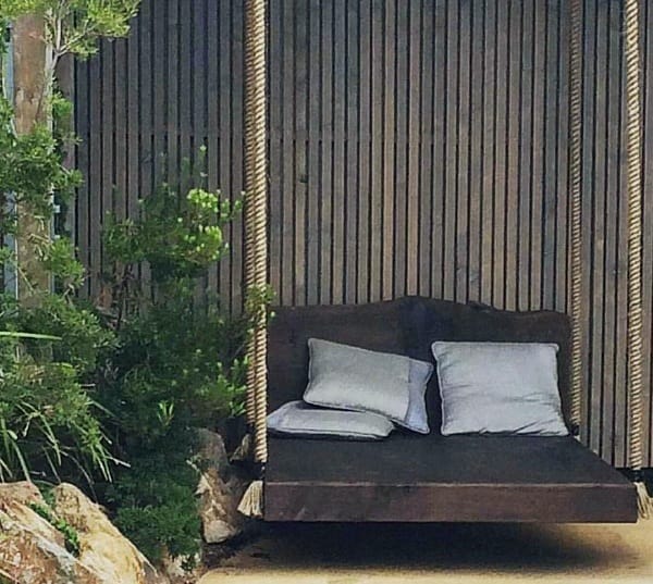 Outdoor hanging bed with thick ropes, dark wooden frame, and cushions surrounded by greenery.