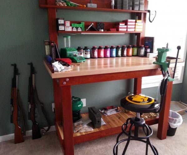 Compact reloading bench with a wooden frame, mounted press, organized shelves for supplies, and nearby rifle storage
