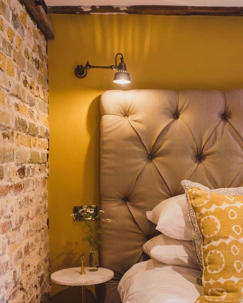 Cozy bedroom corner with a tufted headboard, sunny yellow wall, brick accent, and a small bedside table with a plant