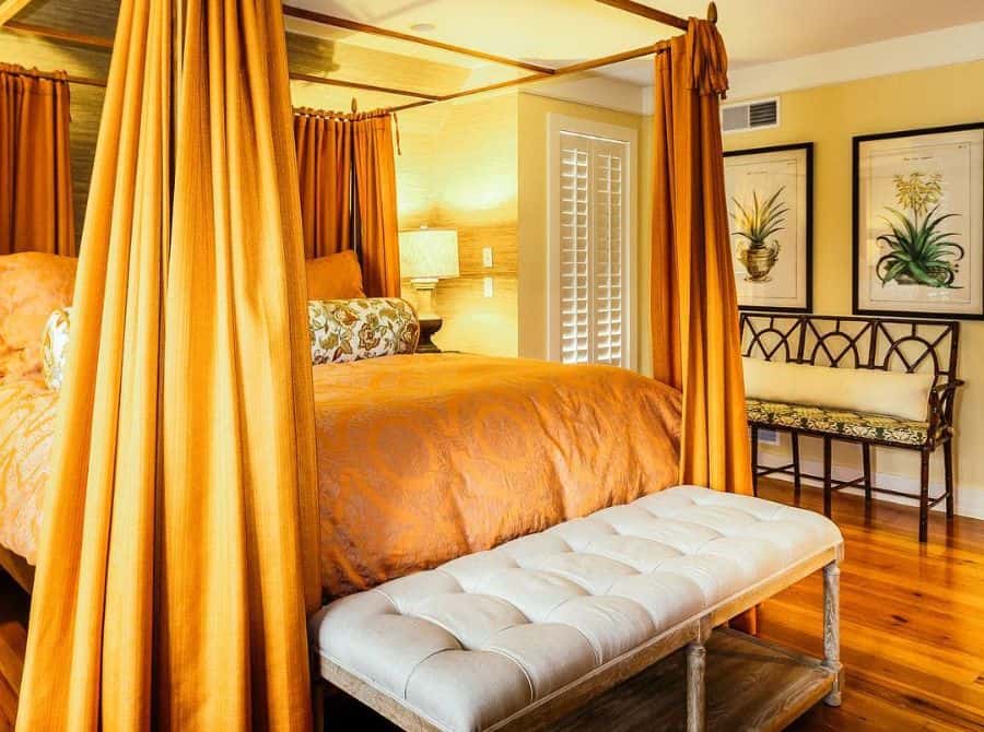 Cozy bedroom with orange canopy bed, yellow accents, tufted bench, and decorative wall art