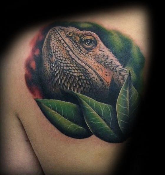 Realistic iguana shoulder tattoo peeking through lush green leaves, with warm earthy tones and a soft shaded background.