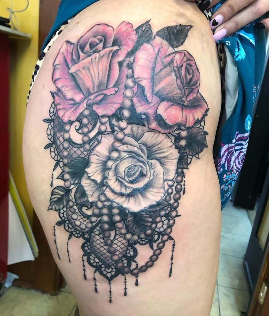 Thigh tattoo featuring vibrant pink and black-and-grey roses intertwined with intricate lace, beaded chains, and ornamental details.