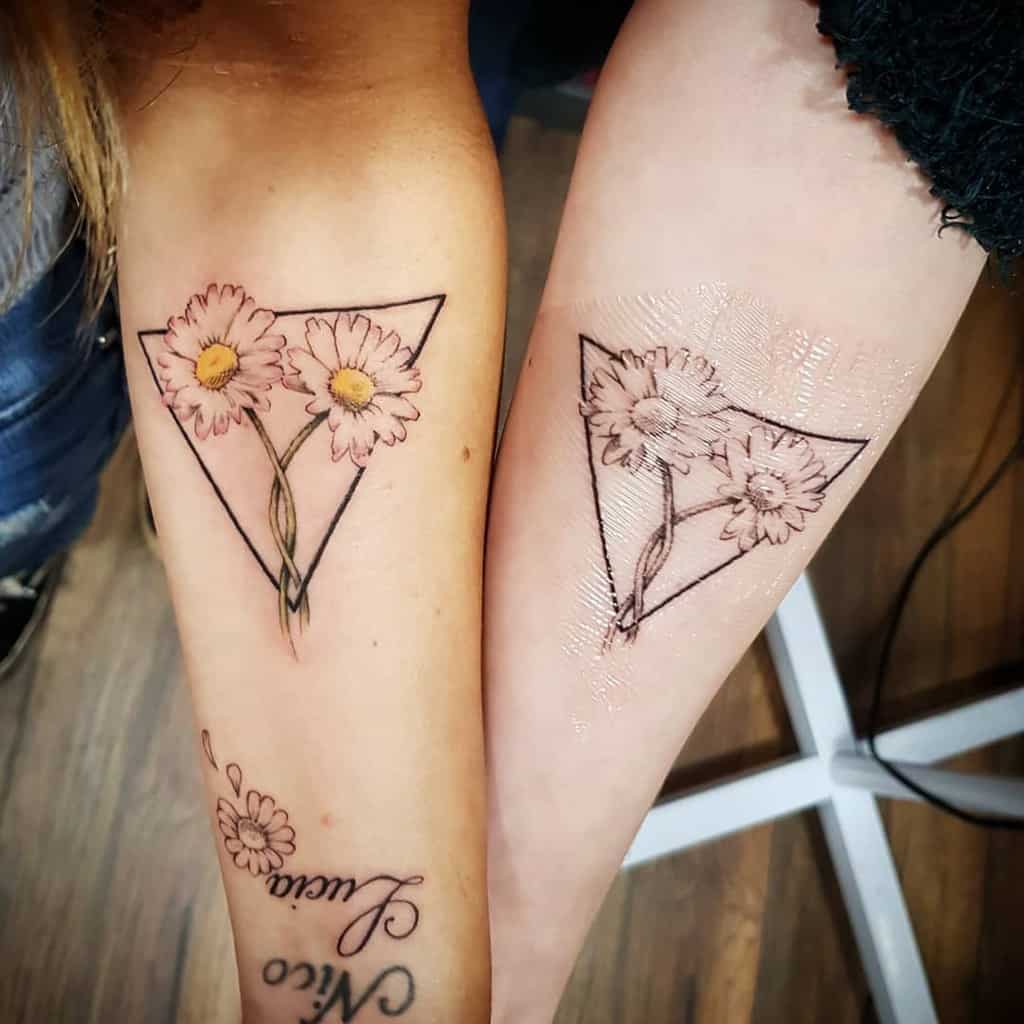 Matching sister tattoos featuring two daisies inside a minimalist triangle outline, symbolizing a close bond