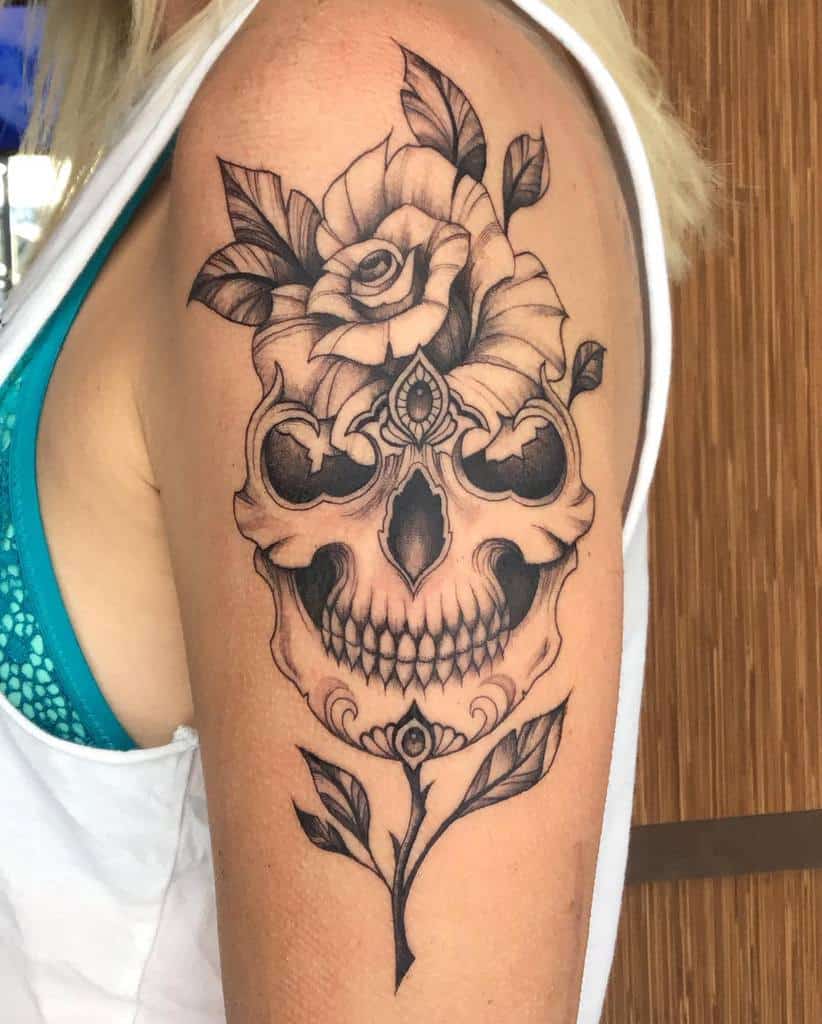 Elegant black ink tattoo of an ornate skull with a blooming rose, intricate linework, and decorative elements on the upper arm.