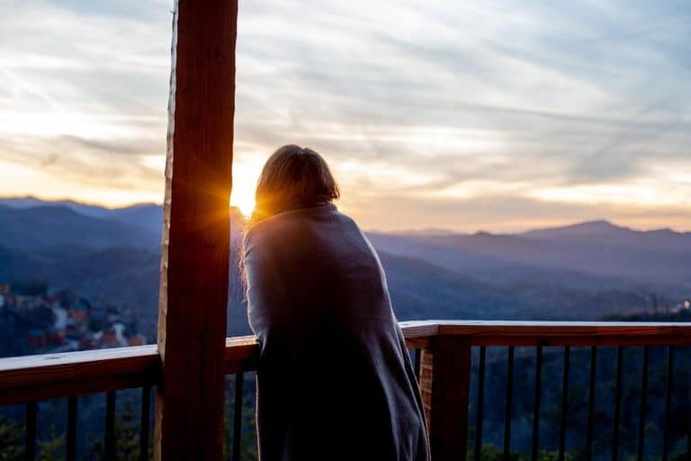 Wrapped in a blanket, a person leans on a wooden balcony at tranquil cabins, watching sunrise over a serene mountain escape