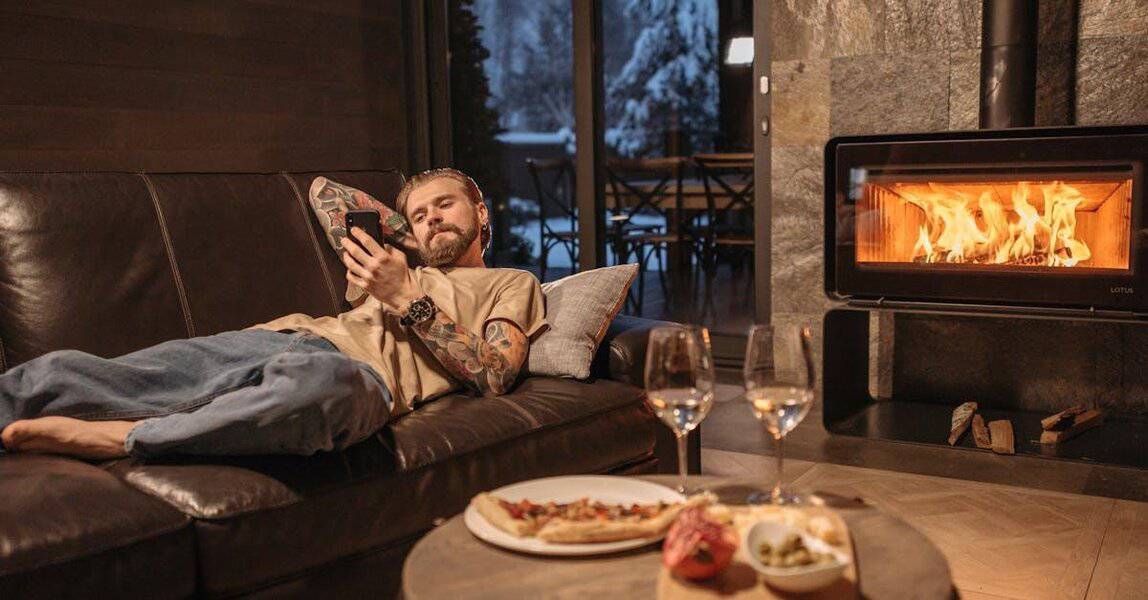 A modern man lounges on a sofa near the fireplace, savoring wine, food, and a magazine in a cozy luxury home