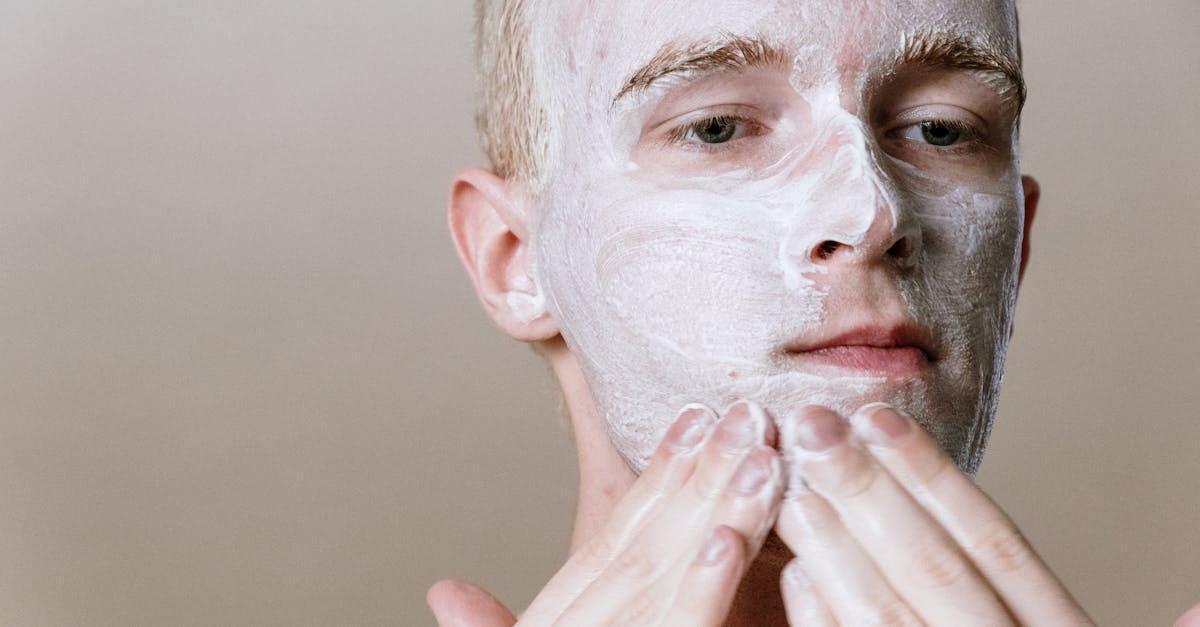 A man uses both hands to put on a white, clinically tested skin care mask, set against a plain background
