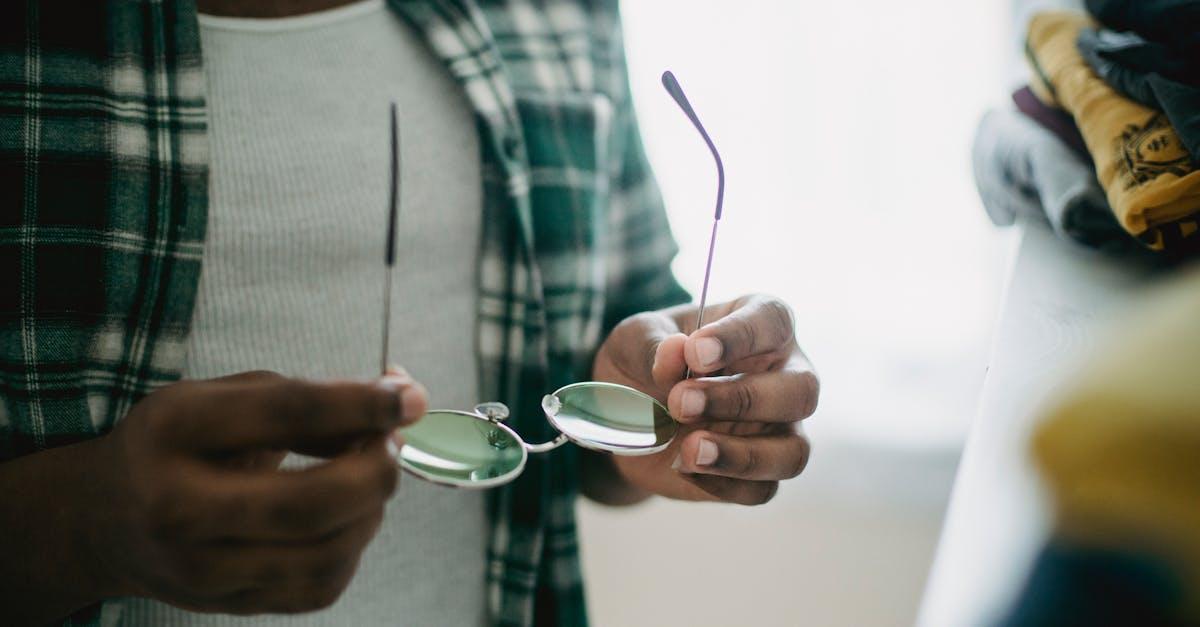 Person in a plaid shirt holding broken eyeglasses—unexpected snag in his EDC essentials near a clothes pile indoors