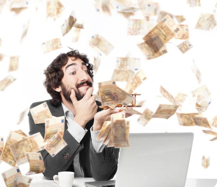 A cheerful man in a suit at a desk with laptop smiles as euro bills fall around him, celebrating higher salaries in Cyprus