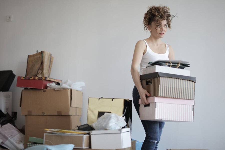 Woman with curly hair in a white tank top carries stacked boxes, surrounded by more boxes and bags in a cluttered room