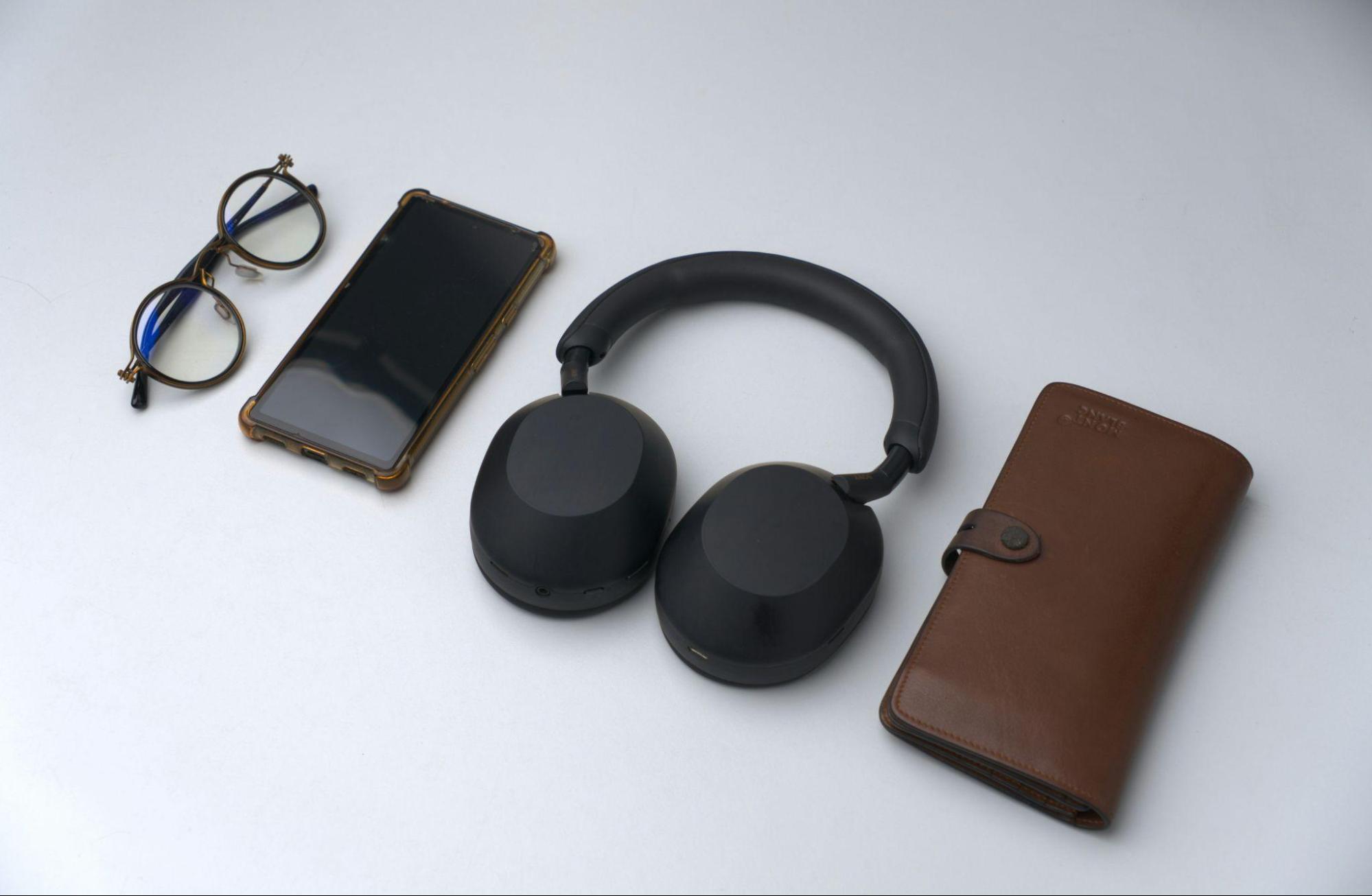 A flat lay of EDC items—eyeglasses, smartphone, black headphones, and a brown wallet—on a light gray background
