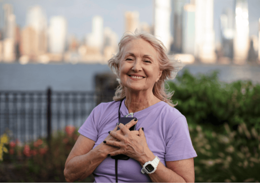 Smiling older woman in purple stands outside holding her phone, confident in citywide mobile coverage and around-the-clock monitoring