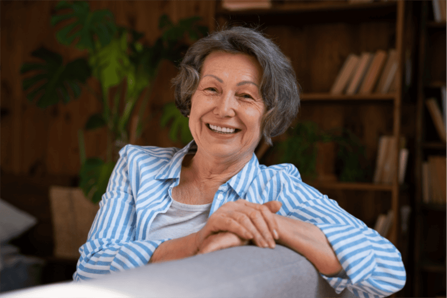 Smiling older woman with gray hair sits on a couch, fall detection device on her wrist and cozy bookshelves behind her