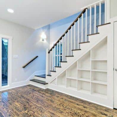 White staircase with built-in under-the-stairs bookshelves and dark wood flooring in a bright room.