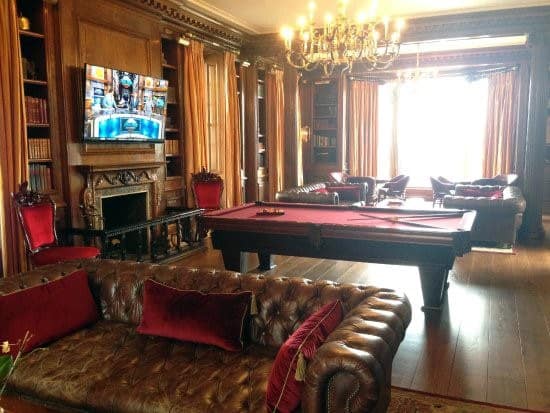 Elegant billiard room with red felt table, leather sofas, chandelier, and wood-paneled walls.