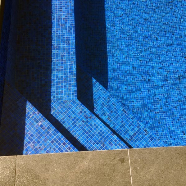 Bright blue mosaic pool tiles with angular shadows and a sleek stone pool deck border.