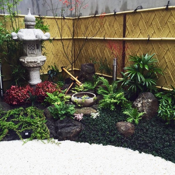 Woven bamboo fence with a diagonal pattern, secured with black ties, in a Japanese garden with a stone lantern, rocks, and plants.