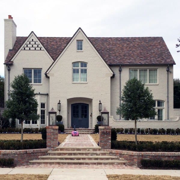 Elegant brick walkway with a staircase leading to a beautiful home, flanked by manicured trees and matching stone pillars