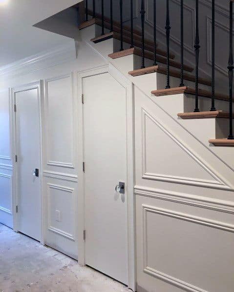 White doors beneath a staircase with decorative paneling, chair rail accents, and dark railings