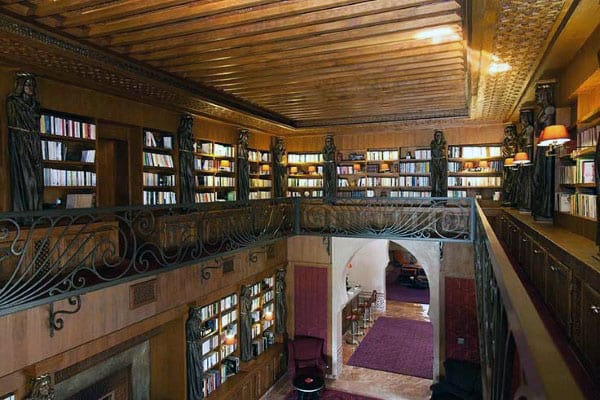 A cozy library with wooden bookshelves, a railing balcony, and statues, softly lit by the warm glow of lamps