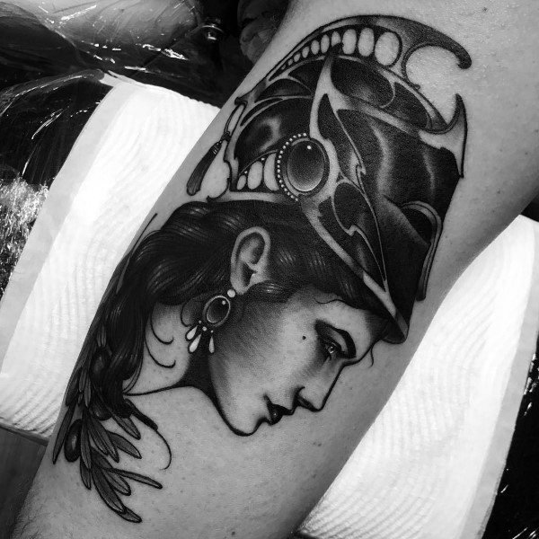 Bold Athena tattoo with a stylized helmet, dramatic shading, and elegant jewelry details in neo-traditional blackwork style on the arm
