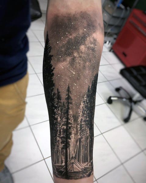 Black and grey forearm tattoo of a starry night sky over a dense forest, featuring a small tent beneath towering pine trees.