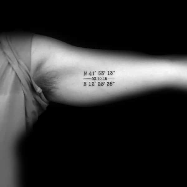 Minimalist coordinate tattoo on the inner bicep with clean black lettering, marking a significant location and date in a subtle design.