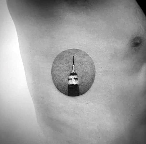 Small Empire State Building tattoo within a shaded circular frame, elegantly inked on a man's ribcage.