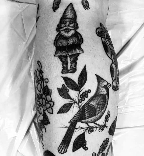 Black and white tattoo of a small gnome with a tall hat and crossed arms, surrounded by floral designs and a detailed bird on a branch.