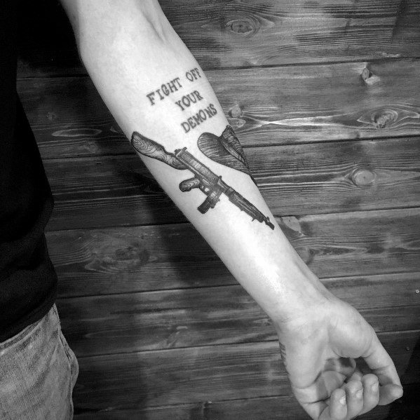 Black and grey Tommy gun tattoo on the forearm, paired with the phrase "Fight off your demons," symbolizing inner battles and resilience.