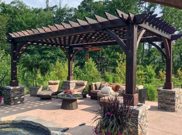 Dark wood pergola with stone pillars, cozy outdoor seating, and a ceiling fan on a paved patio.