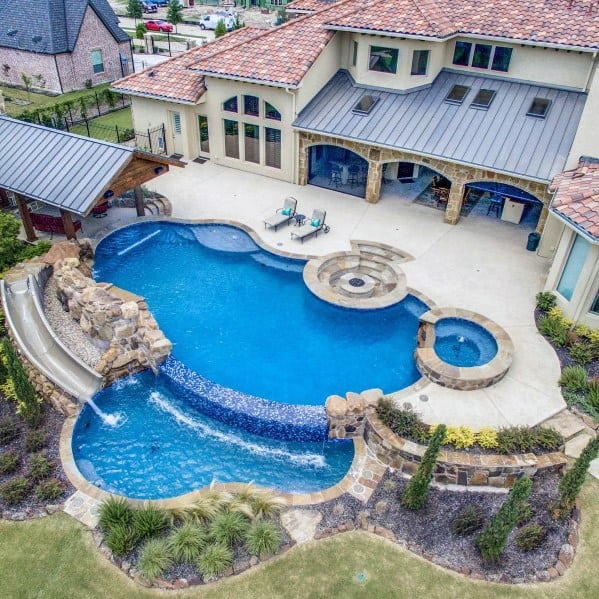 Aerial view of a luxury home with a unique-shaped pool, waterfall, slide, and spa surrounded by landscaping