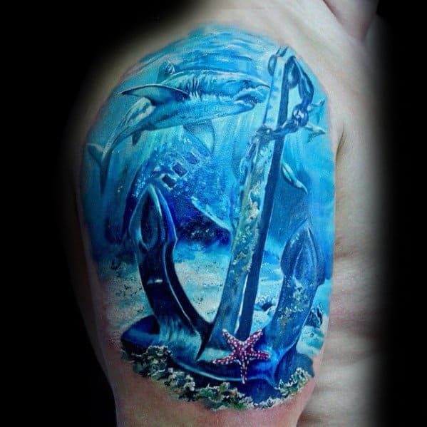 Underwater-themed upper arm tattoo featuring a large anchor, pink starfish, sharks, and a sunken ship in vibrant blue tones.