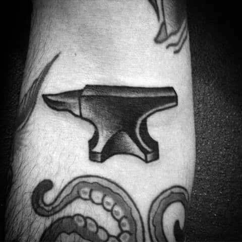 Black anvil tattoo with smooth shading on the arm, placed above tentacle and dagger designs for a bold and balanced composition.