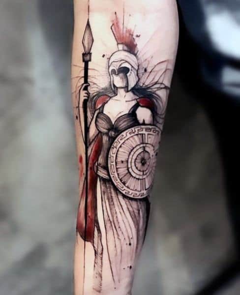 Watercolor-style Athena tattoo with red and black ink accents, featuring a spear, crested helmet, and round Greek shield on forearm