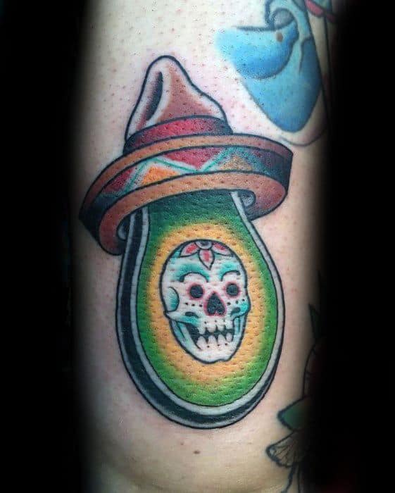 Vibrant avocado tattoo with a sugar skull pit and sombrero hat, blending Mexican cultural elements in a colorful, bold design.