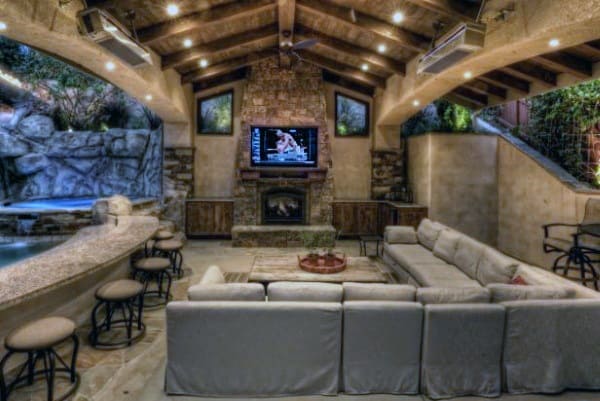 Luxurious poolside pavilion with a stone fireplace, large sectional sofa, bar seating, and mounted TV for entertainment.