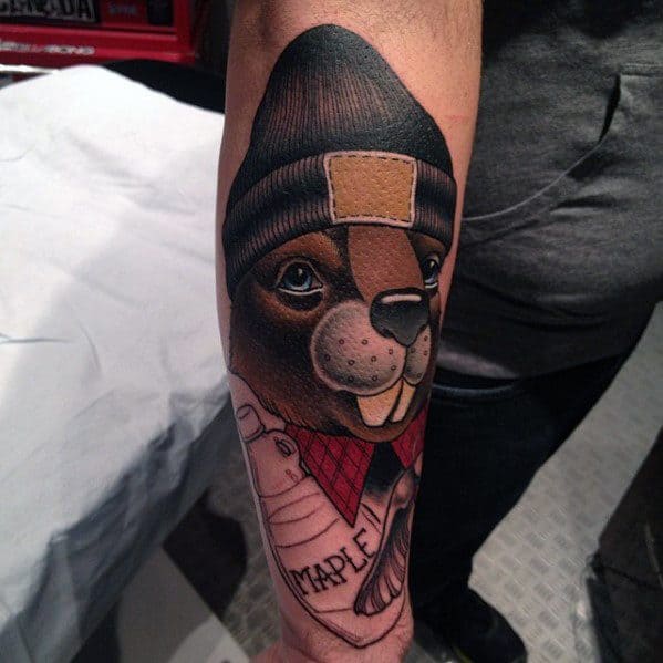 Forearm tattoo of a smiling beaver in a beanie and red plaid scarf, holding a bottle labeled “MAPLE” in a cozy cartoon style.
