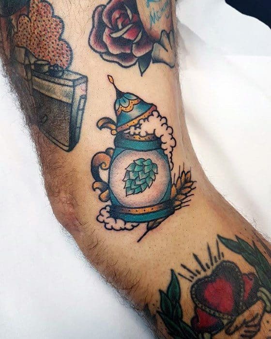 Traditional-style tattoo of a frothy beer stein with a hop cone design on the front, accented by barley and surrounded by bold, colorful outlines.