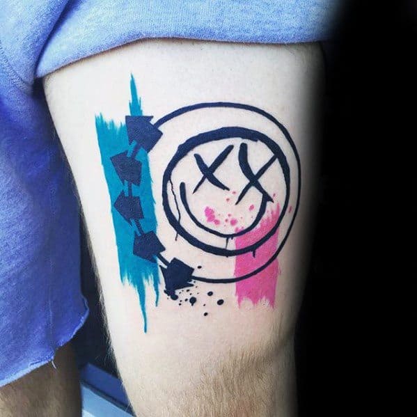 Tattoo of the Blink-182 smiley face logo with black outlines, accented with blue and pink watercolor splashes, creating a vibrant, bold design on the thigh.