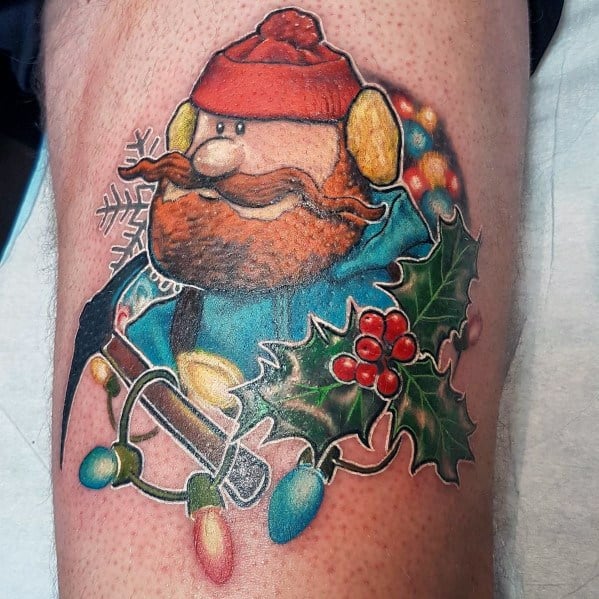 Christmas tattoo of Yukon Cornelius from *Rudolph the Red-Nosed Reindeer*, holding an ice pick, surrounded by holly, festive lights, and a snowflake.