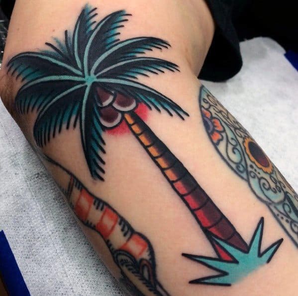 Bold and colorful tattoo of a stylized coconut palm tree with vibrant leaves and coconuts, placed on the upper arm.