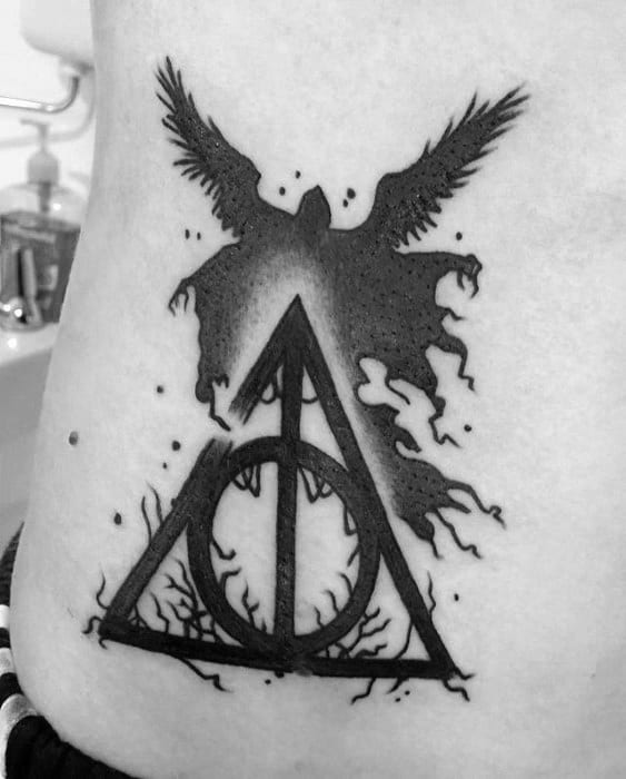 Blackwork tattoo of the Deathly Hallows symbol with a winged Dementor rising from it, surrounded by cracks and shadowy accents.