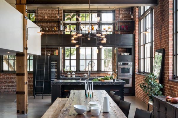 Industrial loft kitchen with exposed brick, large windows, and rustic dining table centerpiece.