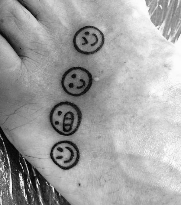 Black and white tattoo of four emoji faces on the top of a foot, each showing different expressions: smiling, grinning, and laughing